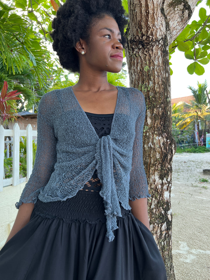 A woman wearing a dusty blue open-knit SHU-SHI shrug cardigan with a front tie, styled over a black top and black skirt, standing outdoors next to a tree. 