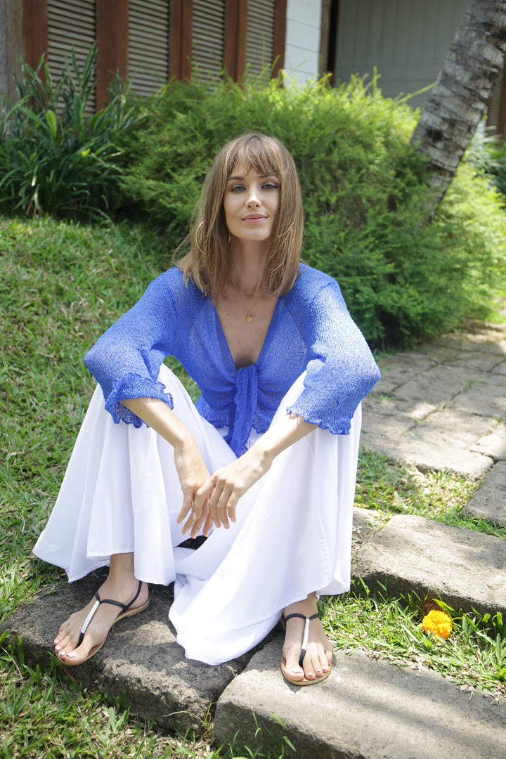 A woman sits outdoors on stone steps wearing a bright blue SHU-SHI Sheer tie-front top and flowing white pants, with greenery and a building in the background.
