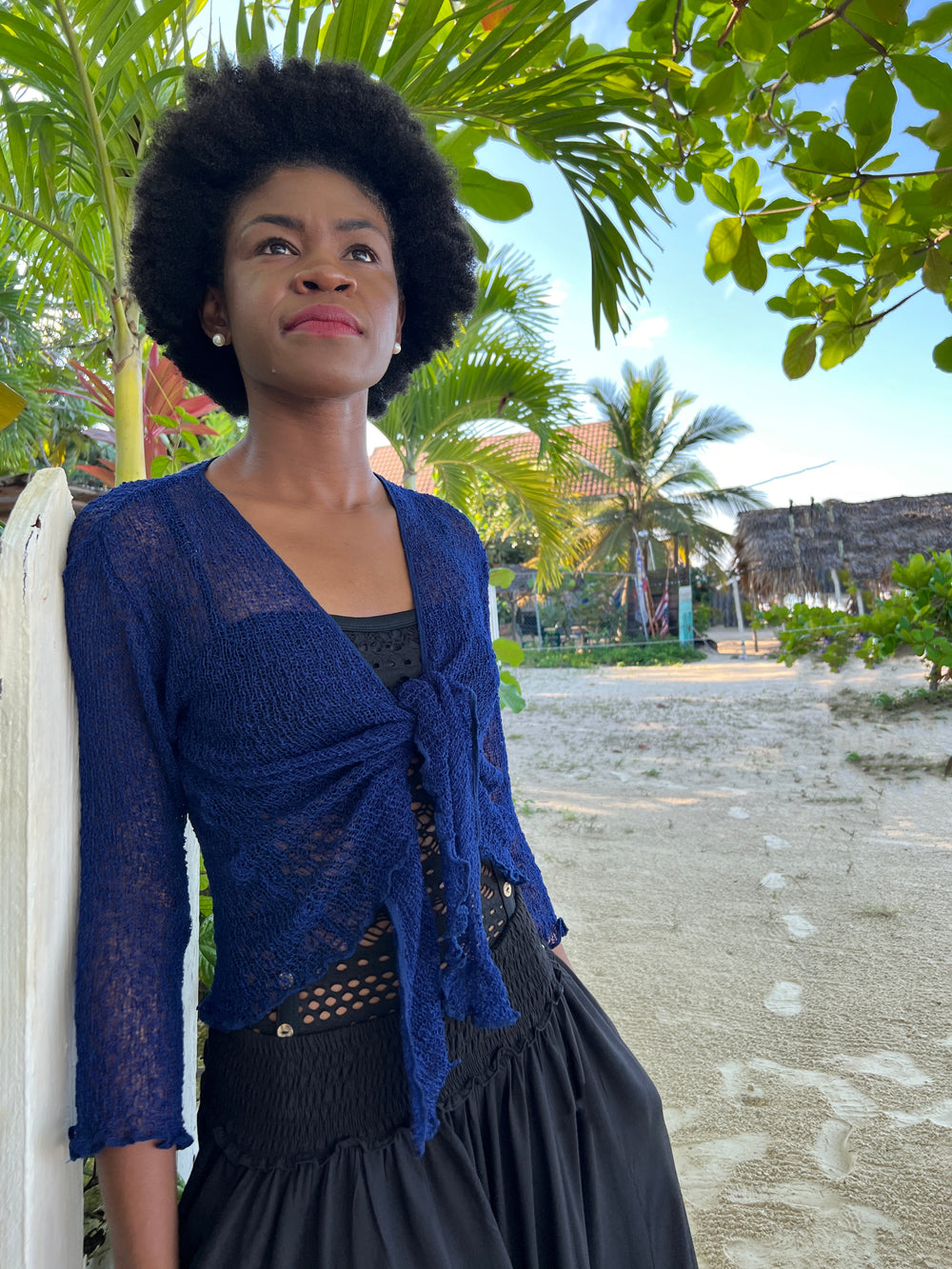 A woman wearing a SHU-SHI dark blue open weave tie front cardigan and black pants, leaning against a white fence in an outdoor tropical setting with palm trees and sand