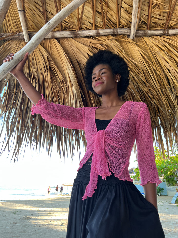 A woman on a beach under a thatched roof wearing a fuchsia SHU-SHI tie front -knit cardigan over a black top and black SHU-SHI pants.