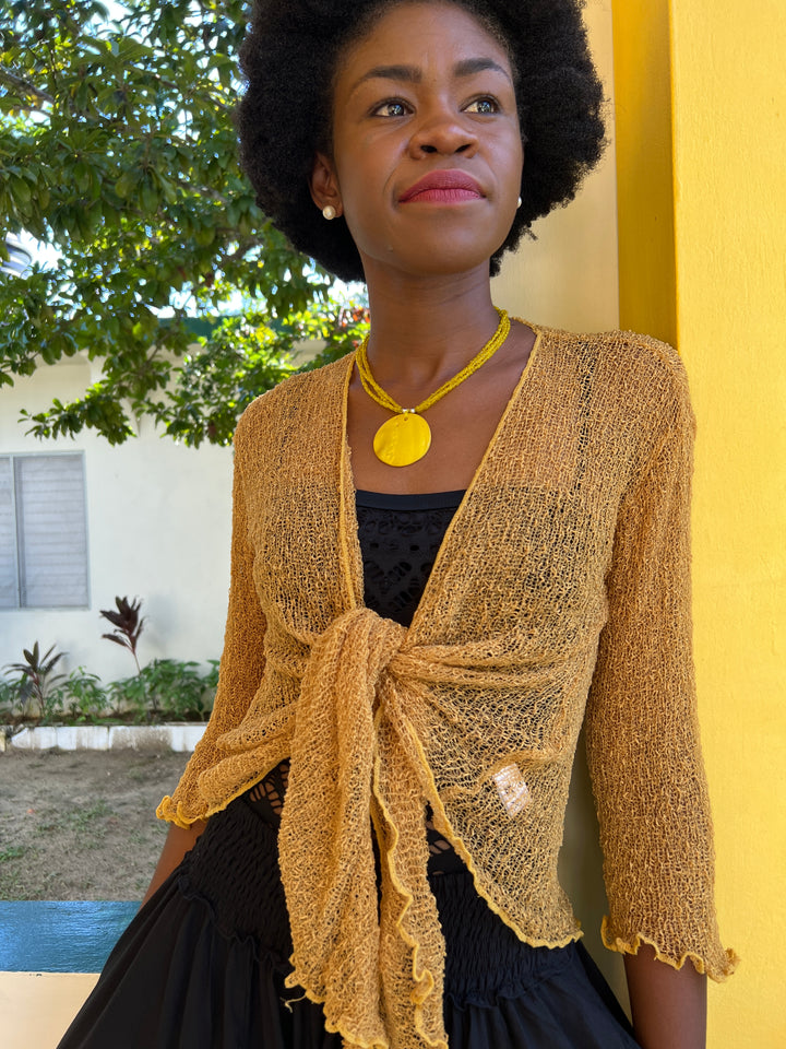 A woman wearing a SHU-SHI gold open-knit cardigan tied at the waist over a black top and black SHU-SHI pants, standing outdoors against a yellow wall.