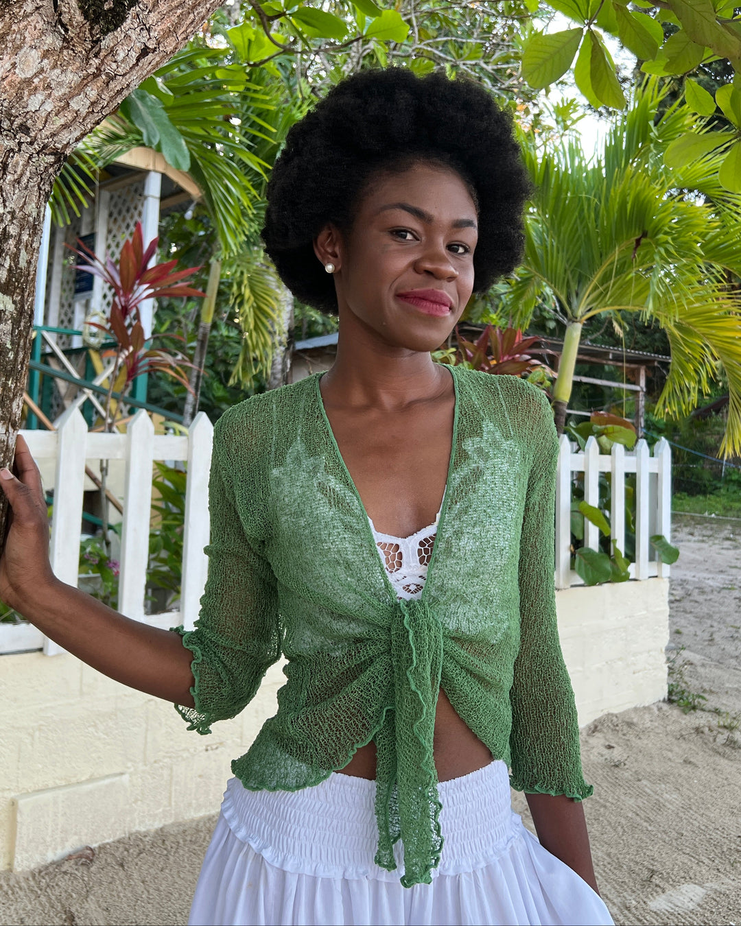 A woman leaning against a tree trunk, wearing a green SHU-SHI open-front knit cardigan tied at the front over a white lace swimsuit and white SHU-SHI Pants. The background shows a tropical beach setting with palm trees, a white picket fence, and sandy ground.