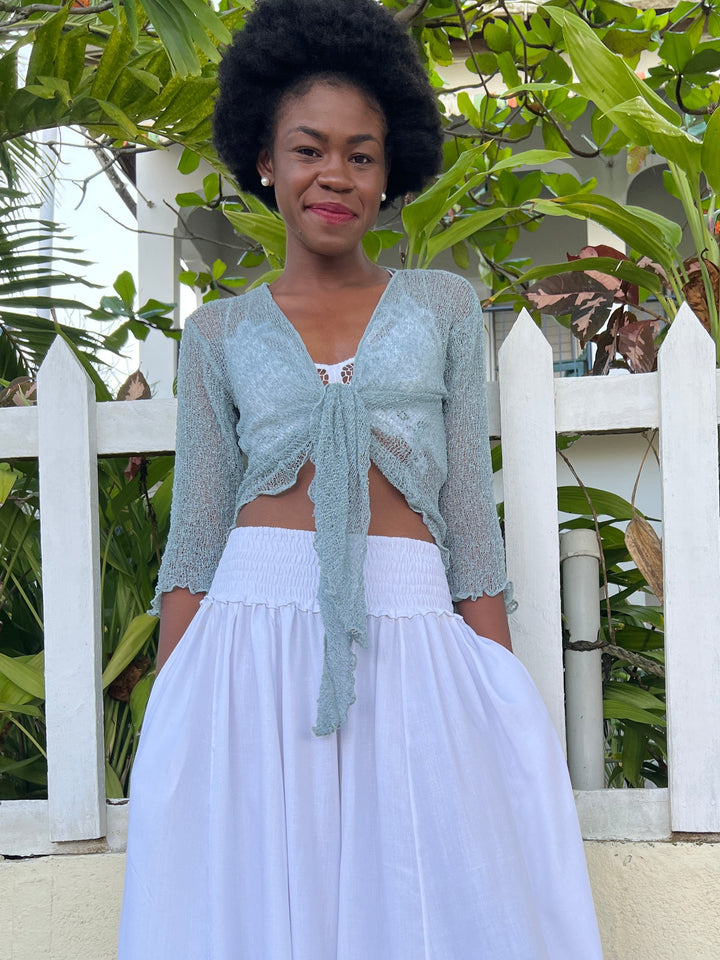 A woman wearing a SHU-SHI grey sheer knit three-quarter sleeve top with a front tie and SHU-SHI white pants, standing outdoors in front of a white picket fence and green plants.