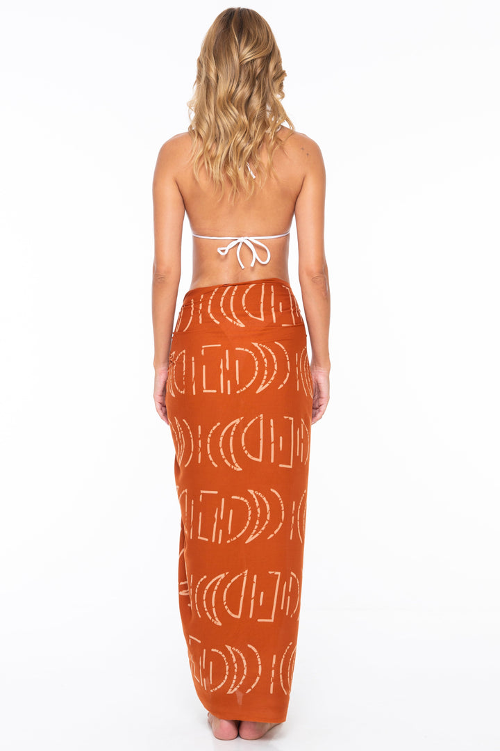 Woman wearing a white bikini top and SHU-SHI moon print sarong in Ginger on a white background - back view